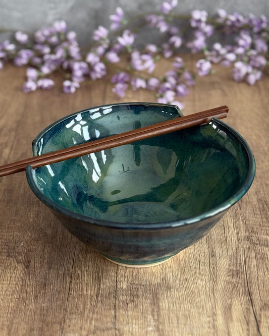 A handcrafted pottery ramen bowl in deep green glaze sits on a wooden surface with chopsticks resting across the cutout rim. Soft purple flowers are blurred in the background.