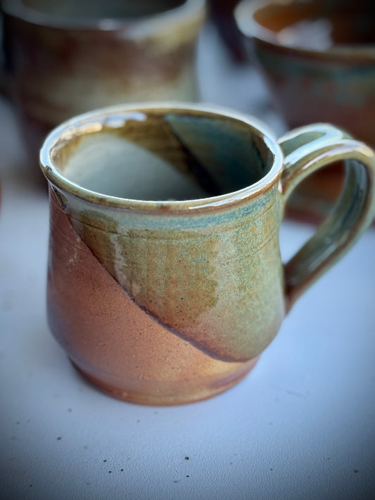 Wood fired ceramic mug with earthy tones of rust, green, and teal, featuring natural ash glaze variation and raw clay texture from the firing process.